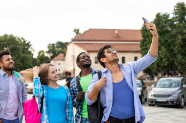 Yetişkin çok ırklı öğrenci grubu akıllı telefon kullanarak selfie çekiyor. Sırt çantalı öğrenciler telefona bakarken gülümsüyorlar.