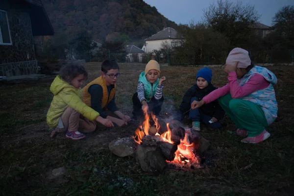 Group of kids in the camp bask by the fire. Schoolchildren play and heat outdoors at autumn