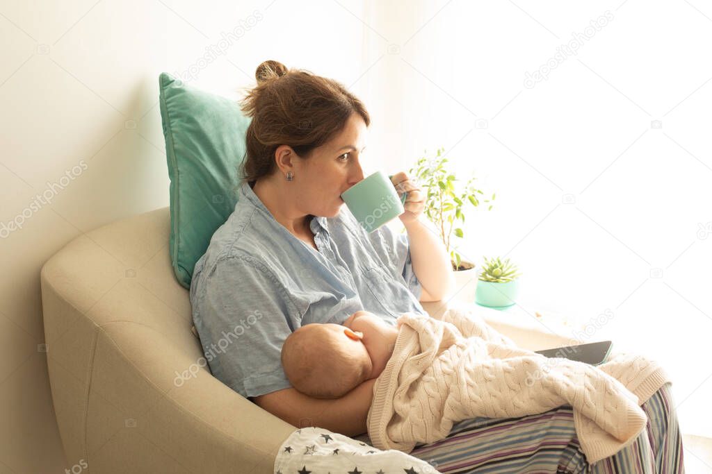 Woman drink tea or coffee during process of breastfeeding of newborn baby. Comfortable breastfeeding