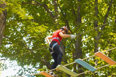 Koruyucu giysili genç kadın asma köprüye basıyor. Koruyucu kıyafetli kadın halat parkında egzersiz yapıyor.