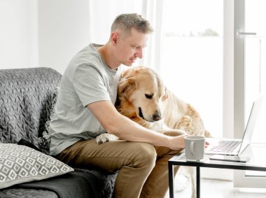 Genç bir adam köpeğiyle birlikte parlak bir oturma odasında bilgisayar başında çalışıyor..