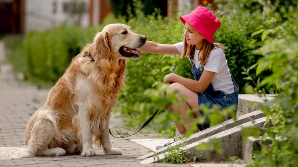 Preteen girl wearing hat with golden retriever dog sitting outdoors in summertime. Pretty kid petting fluffy doggy pet in city