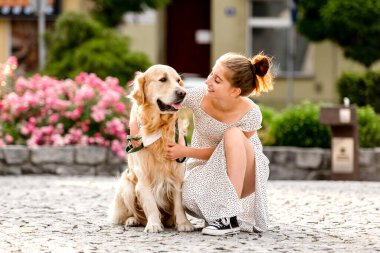 Güzel elbiseli bir kız yazın güneşli bir günde Golden Retriever köpeğine sarılıyor. Şehirde eski mimarisi olan, safkan köpek besleyen güzel bir genç.