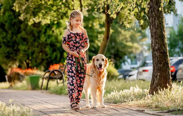 Preteen girl wearing jeans dress with golden retriever dog walking outdoors in summertime. Pretty kid petting fluffy doggy pet in city