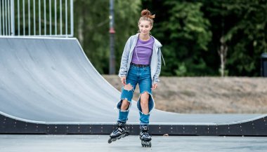 Beautiful girl roller skater riding in city park with ramp. Pretty female teenager rollerskating in casual clothes