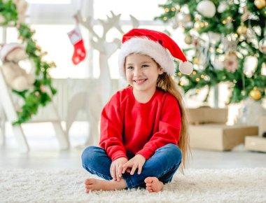 Noel zamanı Noel Baba şapkası takan küçük bir kız Noel ağacıyla süslenmiş evinde oturuyor ve gülümsüyor. Noel zamanı tatlı bir çocuk.