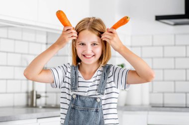Preteen girl with carrots posing at kitchen and smiling. Pretty child with orange vegetables food at home