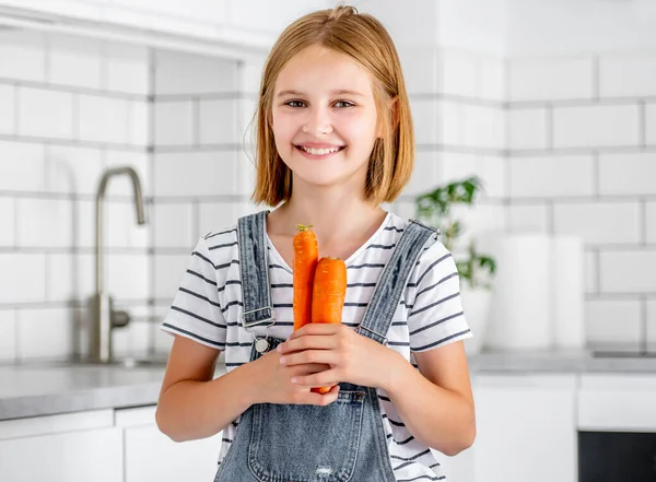 Preteen girl holding carrots at kitchen and smiling. Pretty child with orange vegetables food at home