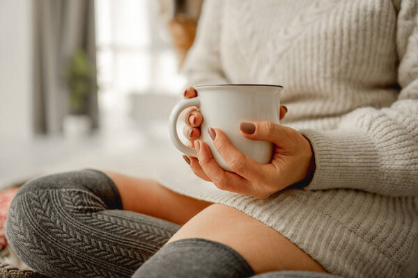 Girl with mug filled with cocoa and marshmallow sitting at home in winter time wearing warm sweater. Cozy portrait of woman with warm beverage in cup