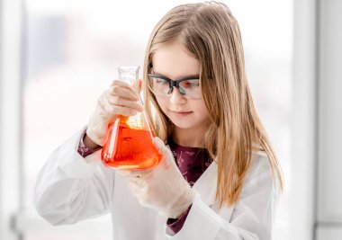 Bilimsel kimya deneyi sırasında koruyucu gözlük takan, elinde turuncu sıvıyla şişe tutan zeki bir kız. Okul fotoğrafı üzerinde kimyasal teçhizatlı kız öğrenci.