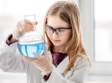 Akıllı kız bilimsel kimya deneyi yapıyor koruyucu gözlük takıyor, şişeyi tutuyor ve mavi sıvıyı ölçüyor. Okul dersinde kimyasal teçhizatlı kız öğrenci.