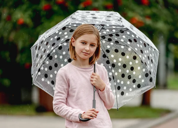 Preteen girl with umbrella looking at camera and smiling outdoors. Pretty child kid portrait in rainy day at street