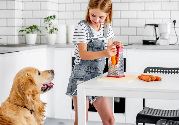 Preteen girl grinds carrot using grater and golden retriever dog looking at child at kitchen. Pretty kid and pet doggy with vegetables at home