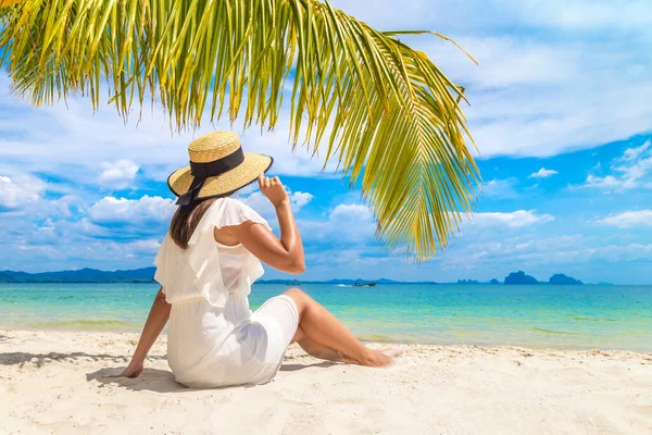 白いドレスと藁帽子を被った若い美しい女性が熱帯のビーチでヤシの葉の下に座っています — ストック写真