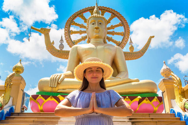 Woman traveler wearing blue dress and straw hat pray at Big Buddha on Koh Samui, Thailand in a summer day