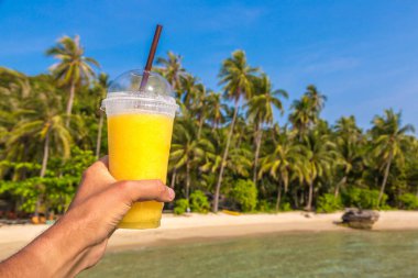 Güneşli bir yaz gününde, tropikal plaj arka planında sarı bir smoothie bardağı tutarken.