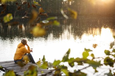 Woman in a yellow jacket relaxing on a wooden pier on a lake with a glass of rose wine watching fall sunset alone. Enjoying nature, relaxation and meditation