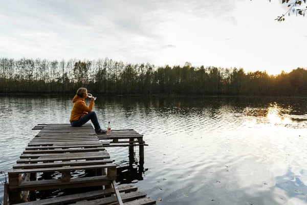 黄色のジャケットを着た女性が湖の上の木製の桟橋でリラックスし 秋の夕日を一人で見ています リラクゼーション 瞑想を楽しむ — ストック写真
