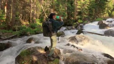 Sırt çantalı bir kadın gezgin, ormanda bir dağ nehrinin yanında su içiyor..