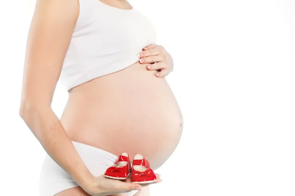 Portrait of Pregnant woman holding pair of red tiny shoes for baby on red background. Studio — Stock Photo, Image