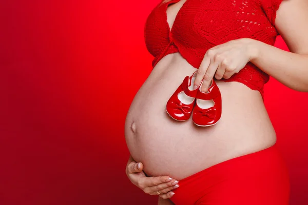 Portrait of Pregnant woman holding pair of red tiny shoes for baby on red background. Studio — Stock Photo, Image