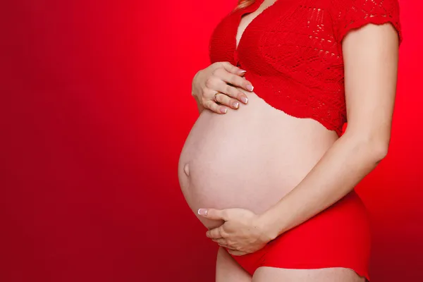 Portrait of Pregnant woman holding pair of red tiny shoes for baby on red background. Studio — Stock Photo, Image