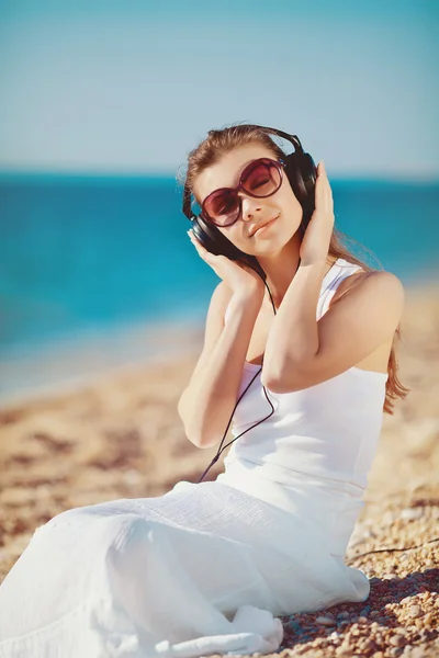 Mooie vrouw luisteren naar muziek tegen blauwe hemel in koptelefoon op de kust — Stockfoto