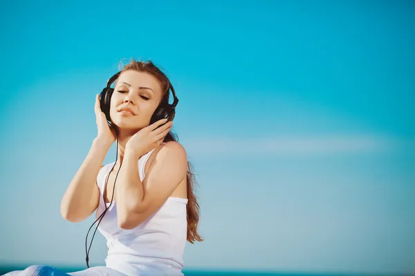 Mooie vrouw luisteren naar muziek tegen blauwe hemel in koptelefoon op de kust — Stockfoto