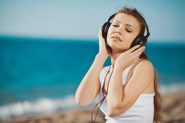 Vacker kvinna lyssna musik mot blå himmel i hörlurar på havsstranden — Stockfoto