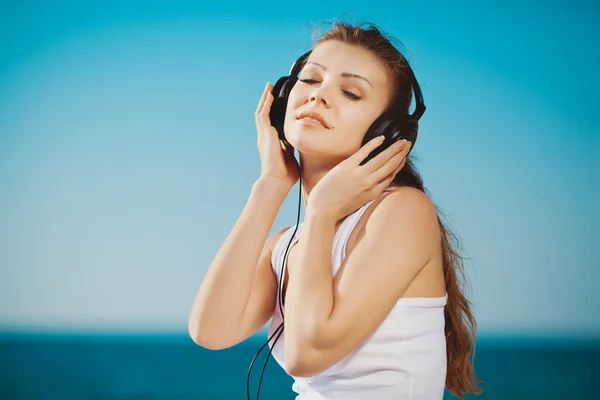 Mooie vrouw luisteren naar muziek tegen blauwe hemel in koptelefoon op de kust — Stockfoto