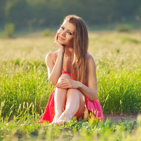 Porträt einer schönen jungen Frau in einem roten Kleid vor einem Hintergrund aus Himmel und Gras im Sommer — Stockfoto