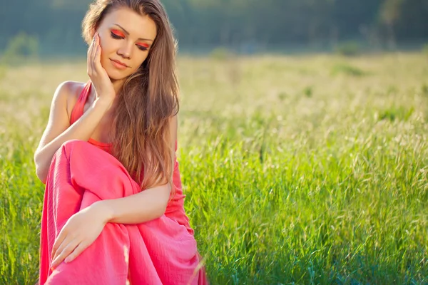 一个美丽的年轻女人，穿着红色的天空背景上，在夏天的草的肖像 — 图库照片