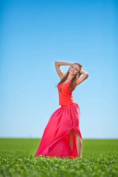 Portrét krásné mladé ženy v červených šatech na pozadí oblohy a trávy v létě — Stock fotografie