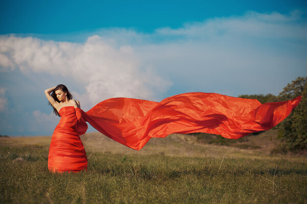 Beautiful young happy woman in long red dress dreams in green field against blue sky