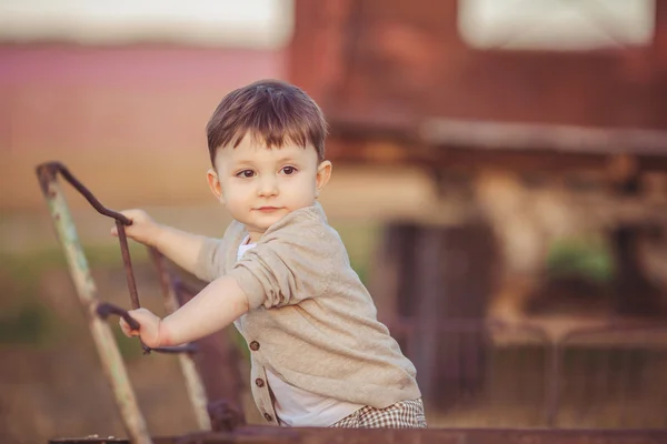 Cute little baby boy standing near metal fence in autumn yard — Stock Photo, Image