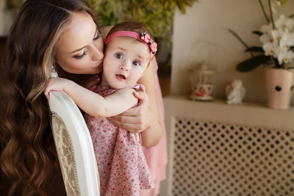 Portret van gelukkige jonge aantrekkelijke moeder spelen met haar babymeisje in de buurt van venster in interieur op haome. roze jurken op moeder en dochter — Stockfoto