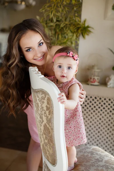 Retrato de feliz joven atractiva madre jugando con su bebé cerca de la ventana en el interior en haome. Los vestidos rosados a la mamá y la hija — Foto de Stock