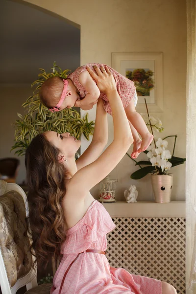 Portrait of happy young attractive mother playing with her baby girl near window in interior at haome. Pink dresses on mother and daughter — Stock Photo, Image