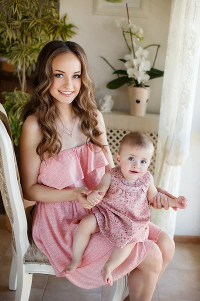 Portrait of happy young attractive mother playing with her baby girl near window in interior at haome. Pink dresses on mother and daughter — Stock Photo, Image