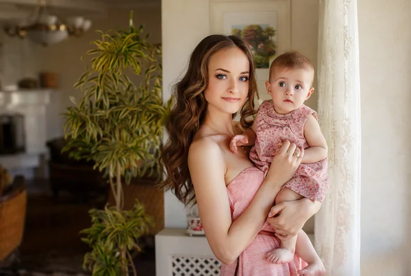 Portrait of happy young attractive mother playing with her baby girl near window in interior at haome. Pink dresses on mother and daughter — Stock Photo, Image