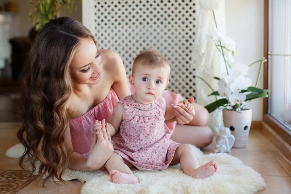 Portrait of happy young attractive mother playing with her baby girl near window in interior at haome. Pink dresses on mother and daughter — Stock Photo, Image