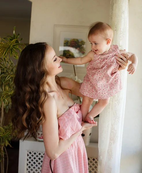 Retrato de feliz joven atractiva madre jugando con su bebé cerca de la ventana en el interior en haome. Los vestidos rosados a la mamá y la hija —  Fotos de Stock