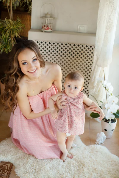 Portrait of happy young attractive mother playing with her baby girl near window in interior at haome. Pink dresses on mother and daughter — Stock Photo, Image