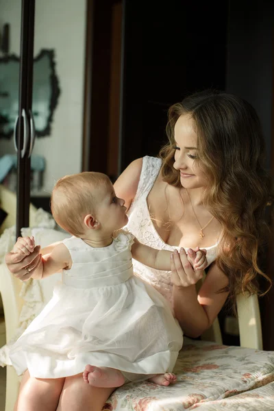 Retrato de feliz joven atractiva madre jugando con su bebé cerca de la ventana en el interior en haome. Los vestidos rosados a la mamá y la hija —  Fotos de Stock