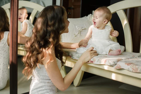 Портрет счастливой молодой привлекательной матери, играющей со своей маленькой девочкой у окна в интерьере дома. Розовые платья на мать и дочь — стоковое фото