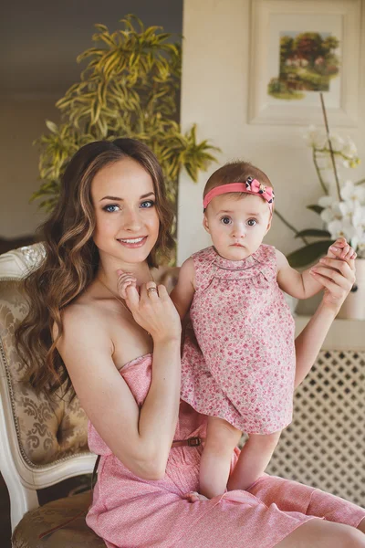 Portrait of happy young attractive mother playing with her baby girl near window in interior at haome. Pink dresses on mother and daughter — Stock Photo, Image