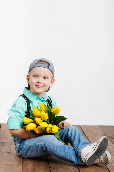 Smiling boy holding a bouquet of yellow tulips in hands sitting on wooden floor — Stock Photo, Image