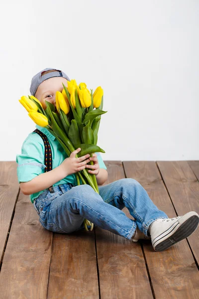 Sonriente niño sosteniendo un ramo de tulipanes amarillos en las manos sentado en el suelo de madera — Foto de Stock