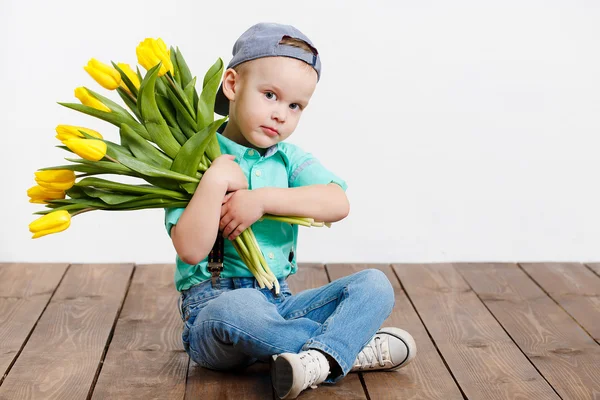Leende pojke håller en bukett med gula tulpaner i händer sitter på trägolv — Stockfoto