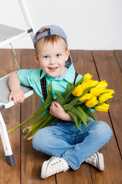 Sonriente niño sosteniendo un ramo de tulipanes amarillos en las manos sentado en el suelo de madera — Foto de Stock